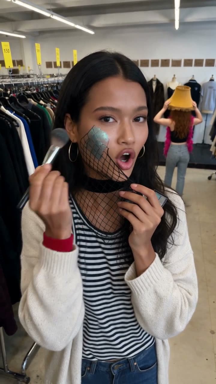 Marguerite dabbing shimmer through fishnet on her cheek in a thrift store aisle, a frame from the fifth take of clip two